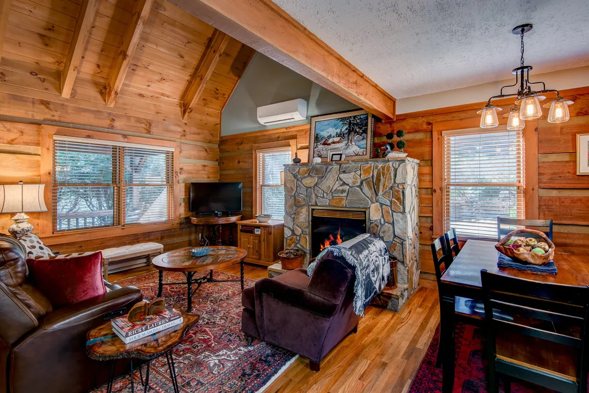 This interior image showcases a stone fireplace as a central focal point, complemented by striking wooden beams overhead. The scene includes a dining table and comfortable seating, emphasizing the open-plan nature of the living area. Warm lighting and cozy decor highlight the inviting atmosphere, perfect for relaxing by the fire or gathering for a meal.