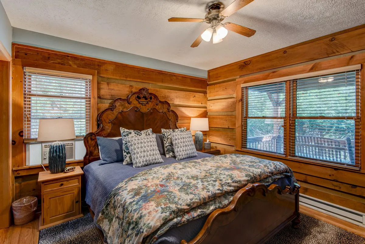 A sunlit room with a wooden bed frame, floral bedding, and multiple windows allowing natural light to filter in. The space feels airy and serene, inviting relaxation and quiet mornings.
