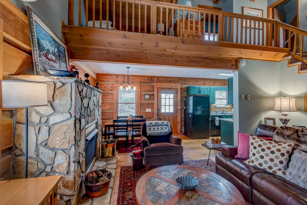 The image presents a welcoming open-plan living area with a stone fireplace, exposed wooden beams, a plush sofa adorned with pillows, a dining table, and a glimpse of the kitchen area. The hardwood floors and stylish furnishings add an elevated, luxurious touch, while the arrangement invites relaxation and socialization.