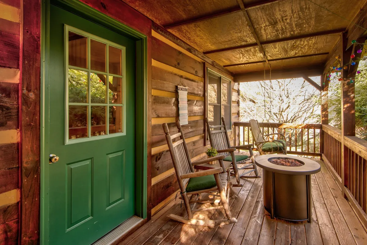 The image shows the welcoming front porch of the cabin, featuring inviting green doors, classic rocking chairs, and a cozy fire pit. The rustic wooden walls and leafy surroundings set the stage for outdoor relaxation, sunset watching, or enjoying a glass of wine together after a day of exploring.