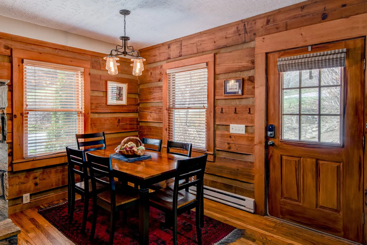 A cozy dining area with a wooden dining table, black chairs, rustic walls, a charming light fixture, and a decorative bowl of fruit, evoking warmth and a welcoming ambiance for family dinners or game nights.