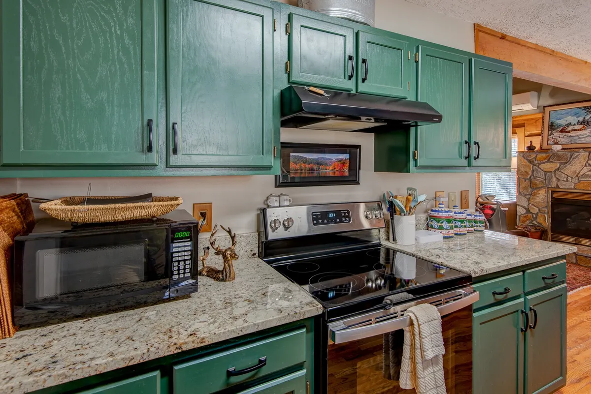 A warm, inviting view of the kitchen featuring green cabinets, granite countertops, stainless steel appliances, and decorative items on the shelves. The setup showcases ample workspace and a cozy, functional design ideal for cooking and socializing.