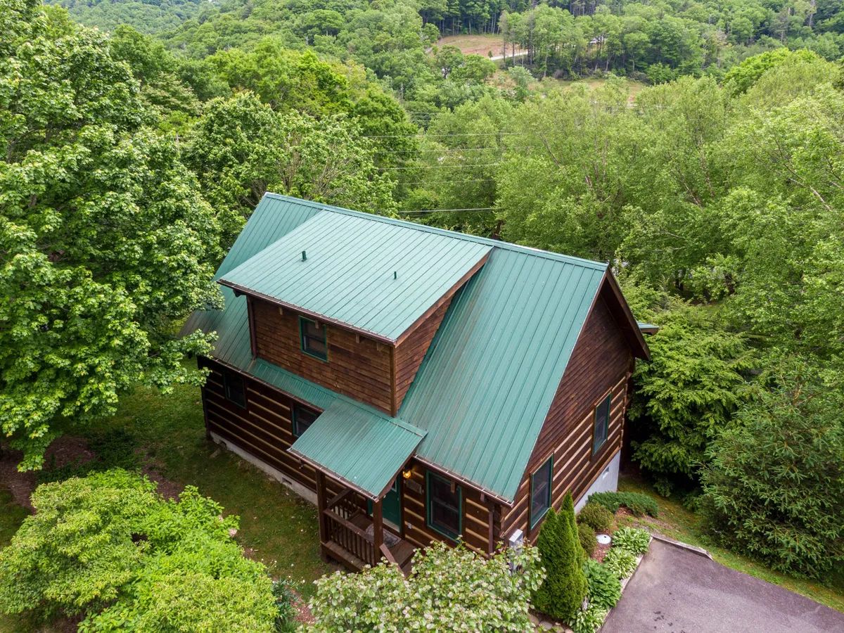 A log cabin with a green metal roof nestled among tall trees, surrounded by a landscaped garden and open green space. The porch and garden area suggest ample room for outdoor activities and gatherings.