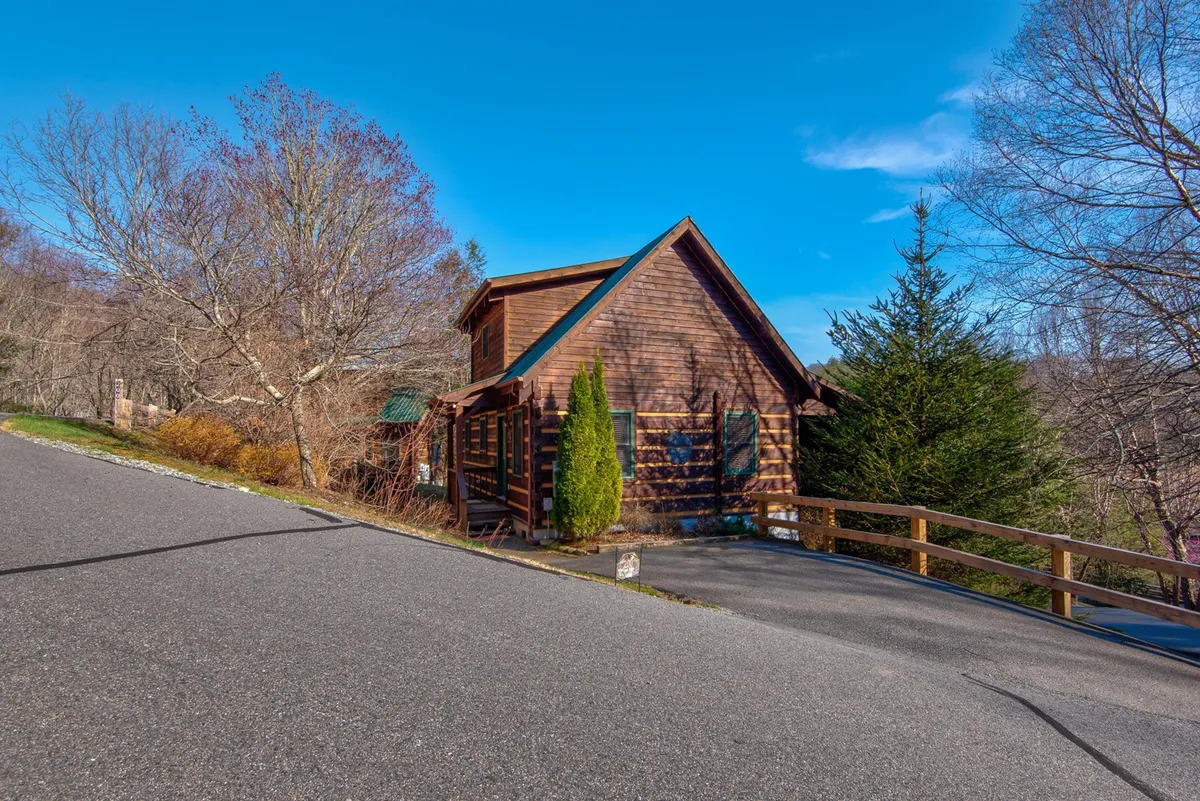 The image showcases the exterior of a charming log cabin surrounded by lush greenery, with a welcoming driveway leading up to the front porch. The peaceful setting hints at opportunities for outdoor relaxation, family gatherings, and enjoying nature right outside the door.