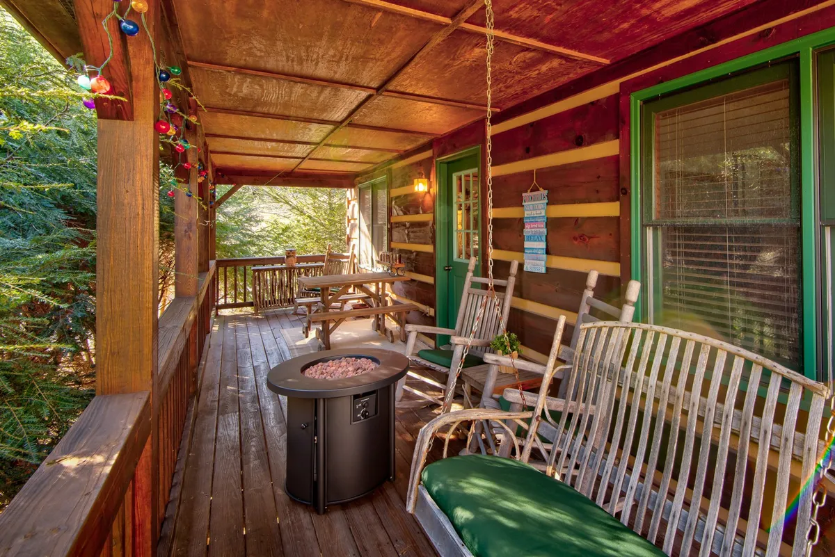 A cozy exterior porch scene with wooden rocking chairs, a fire pit table, a wooden table, and decorative lights strung above. The porch is surrounded by natural greenery and offers plenty of space for relaxation, dining, and socializing outdoors.