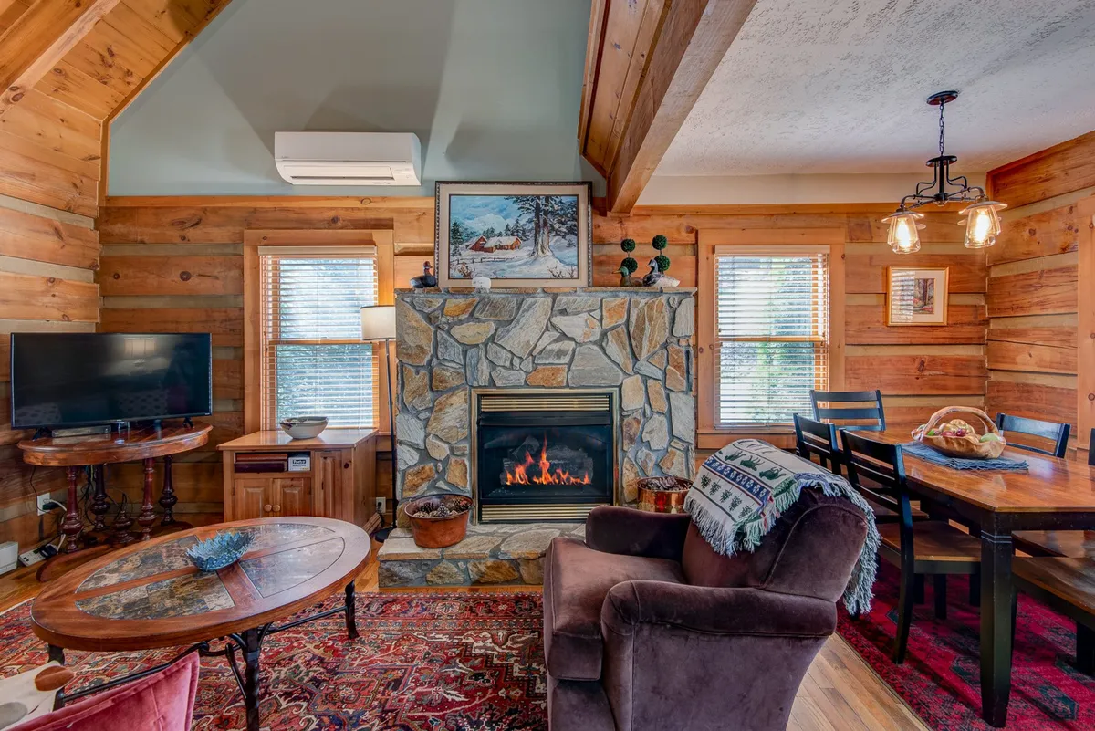 The interior living area of the cabin, featuring a cozy stone fireplace, comfortable seating, a dining table, and a TV. The space is bathed in warm light, creating an inviting atmosphere perfect for family movie nights or board games after a day out.