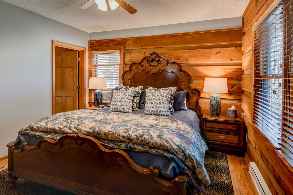 A cozy bedroom featuring an ornate wooden headboard, floral bedding, matching lamps on nightstands, and rustic wooden paneling. Sunlight streams softly through the window, bathing the space in a tranquil glow.