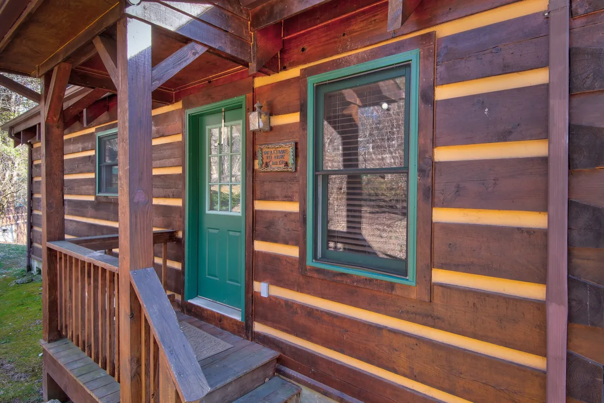 A cozy exterior entrance view featuring a green door, wooden steps, and log siding. The inviting porch is ideal for relaxing with pets after outdoor adventures, evoking the welcoming spirit of the retreat.