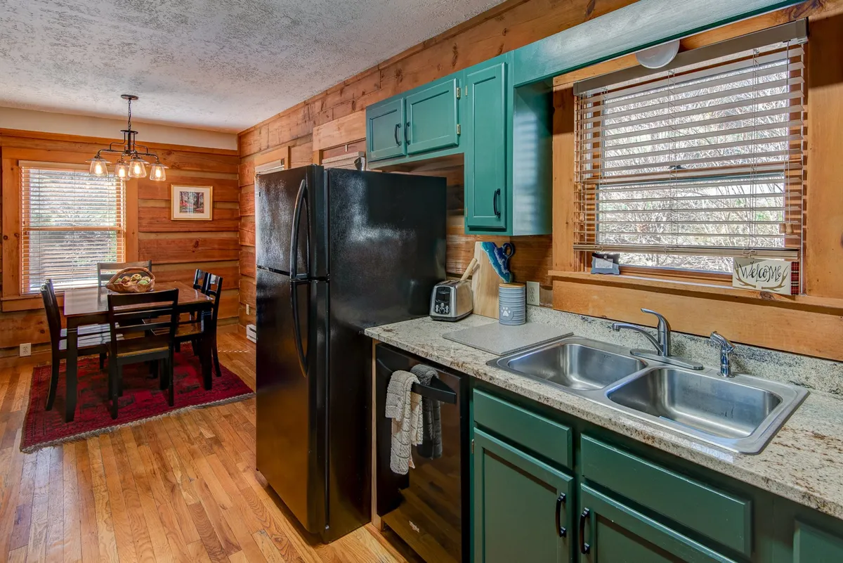 A welcoming space that combines the kitchen and dining area. The image reveals green cabinetry, a black refrigerator, a rustic dining table, and wooden walls, establishing a seamless flow between cooking and dining zones.
