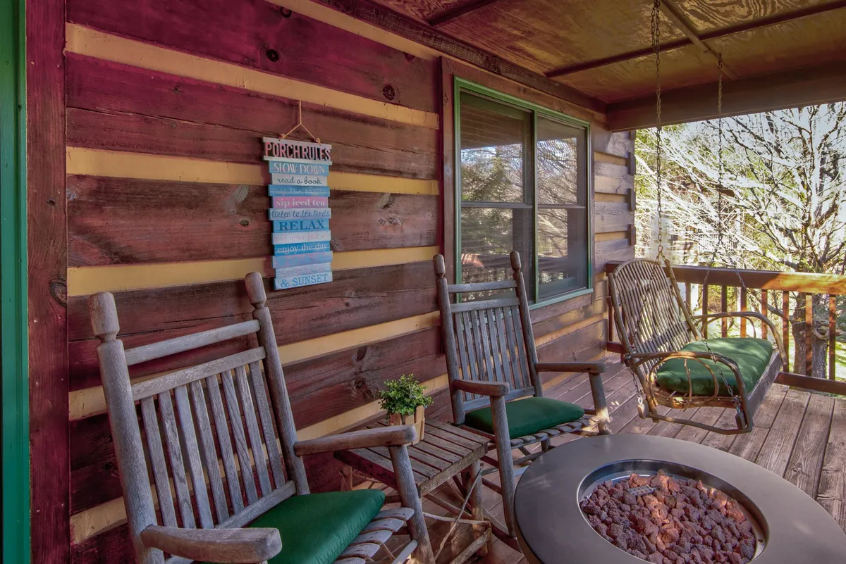 An exterior scene centered around a pair of rocking chairs by a fire pit, accompanied by a decorative sign. The setup encourages socializing, enjoying the fire’s warmth, and relaxing under open skies, with the porch seamlessly connecting to nature.