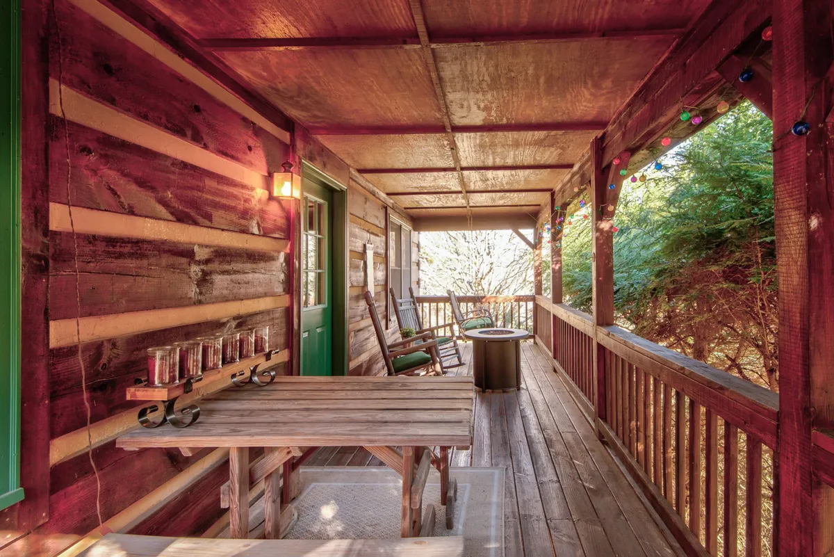 A welcoming exterior view of the porch featuring a wooden table, green door, rocking chairs, and string lights. The porch exudes a cozy, inviting ambiance, perfect for lounging, entertaining, or sharing quiet moments with pets and loved ones.