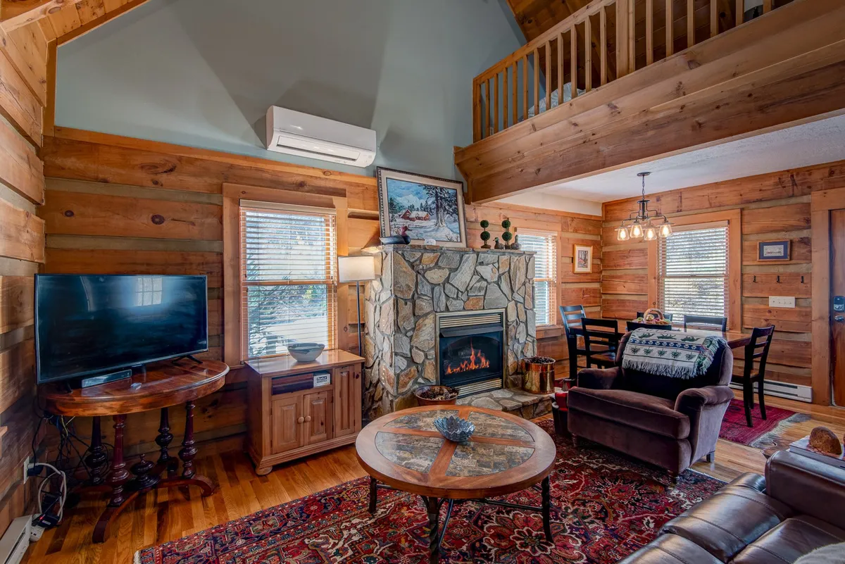This cozy interior scene centers around a stone fireplace, complemented by wooden furniture, a warm rug, and artwork on the walls. The room radiates a welcoming mood, perfect for snuggling up with a blanket or enjoying a family game night.